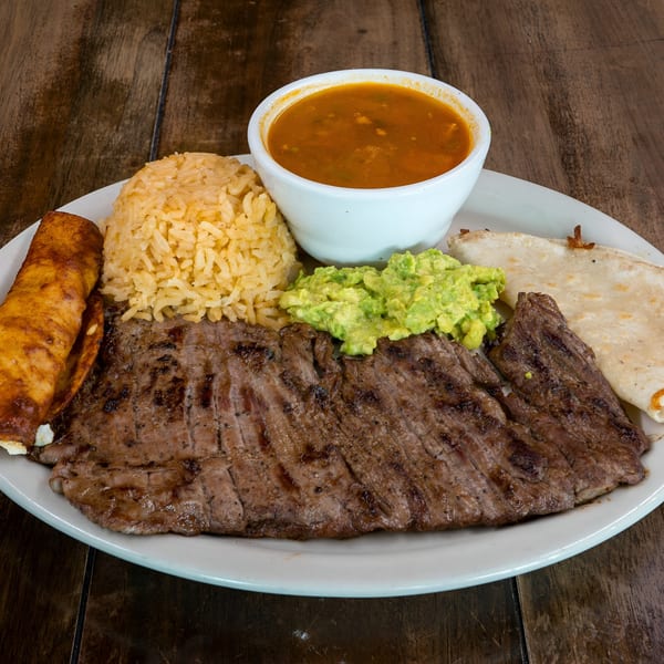 Grilled steak, rice, guacamole, a tortilla, and a bowl of soup on a white plate.