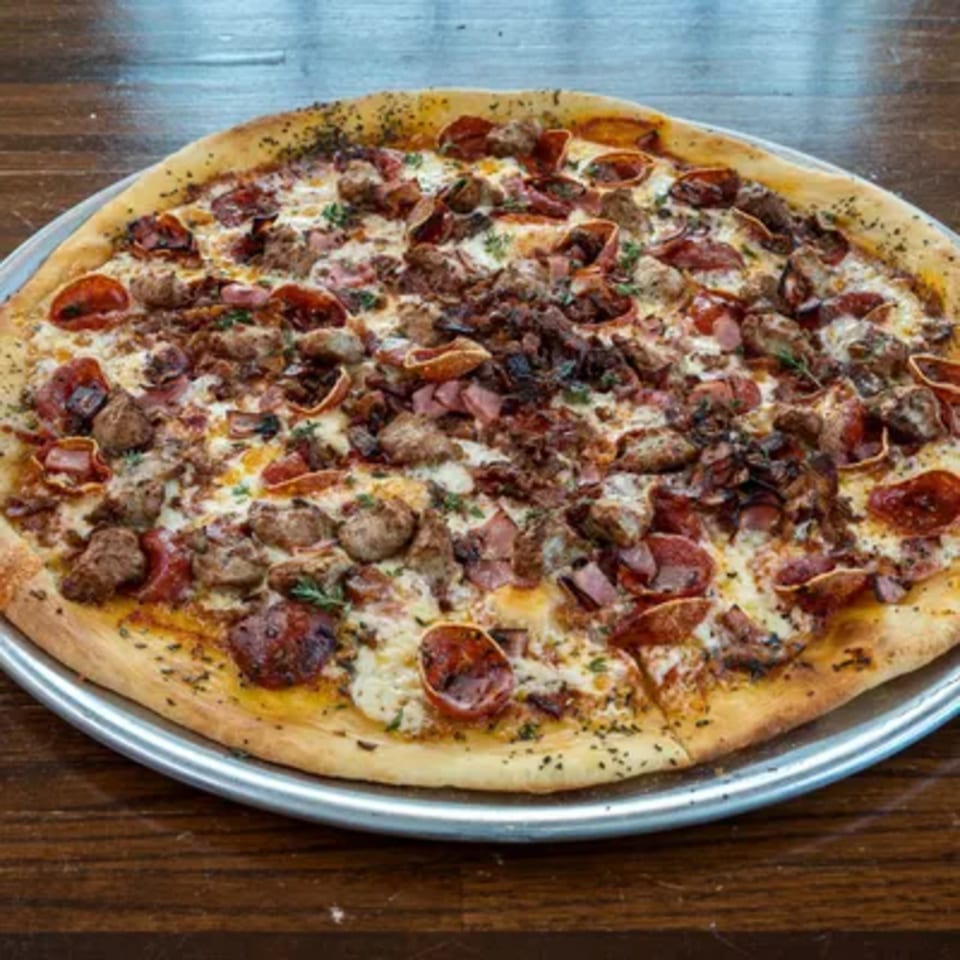 A large, round Blue Eye Bistro meat lover's pizza with pepperoni, sausage, and ham on a metal tray.