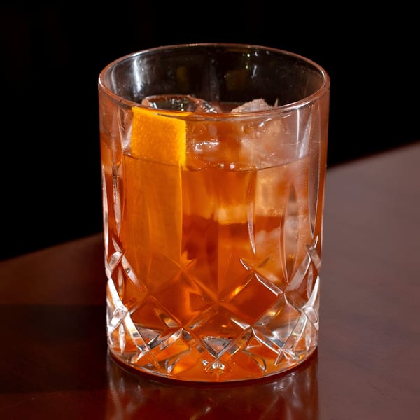 An Old Fashioned cocktail with an orange peel and ice in a cut glass on a dark table.