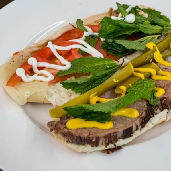A Blue Eye Bistro sandwich with sliced meat, tomatoes, cheese, pickles, mint, and sauces on a white plate.