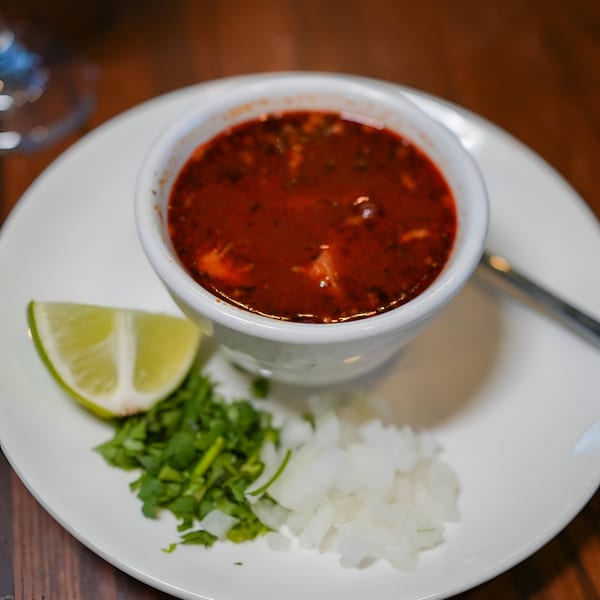 A bowl of red soup with a lime wedge, chopped cilantro, and diced onions on a white plate.