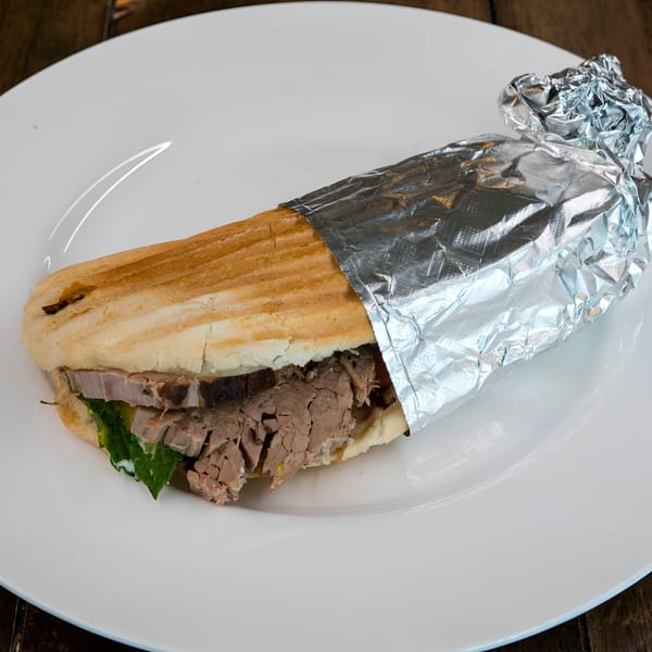 A Blue Eye Bistro panini with sliced meat and lettuce, partially wrapped in foil on a white plate.