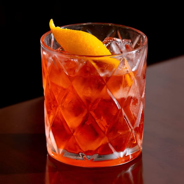 A vibrant red cocktail with ice and an orange peel garnish in a textured glass on a dark table.