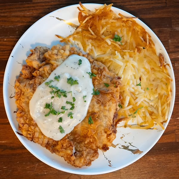 Crispy fried chicken steak with creamy gravy and hash browns on a white plate.